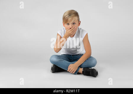 Piccolo ragazzo biondo si siede con le gambe incrociate sul pavimento ed è sorpreso che copre la bocca Foto Stock