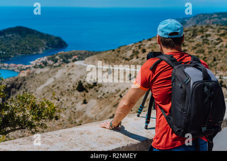 Maschio vacanza estiva vacanza sull'isola di Cefalonia in Grecia. Fotografo con zaino godendo di cattura del villaggio mediterraneo Assos dall'alto punto di vista. La fotocamera sul cavalletto Foto Stock