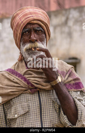 Angadihalli, Karnataka, India - 2 Novembre 2013: Ritratto di uomo anziano con il bianco e il rosso barba di fumare una sigaretta beedi. Indossa turbante, a scacchi sh Foto Stock