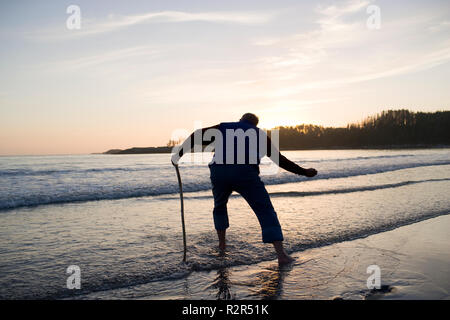 Adulto Uomo scrematura rocce al bordo dell'acqua su una spiaggia al tramonto. Foto Stock