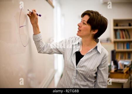 Metà femmina adulta insegnante scrivere sulla lavagna all'interno di una classe. Foto Stock