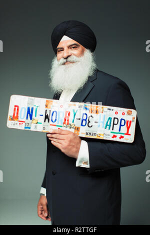 Ritratto sorridente, ben vestito uomo senior in turbante holding non preoccupatevi di essere felice di targhe Foto Stock