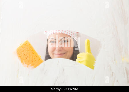 Donna che indossa guanti gialli e bandana azienda spugna arancione in una mano con il pollice fino a sorridere da dietro il vetro doccia Foto Stock