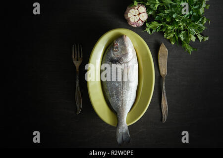Vista dall'alto di uno crudo fresco dorada il pesce in un piatto verde con prezzemolo e aglio, forchetta e coltello su una tavola nera Foto Stock