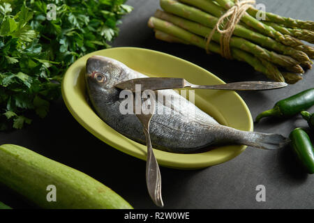 Un crudo fresco dorada il pesce in un piatto verde con verdure verdi e posate su una tavola nera Foto Stock