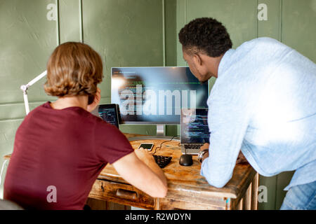 Due giovani programmatori che lavorano con il codice di un programma seduta sul posto di lavoro con tre monitor in ufficio sulla parete verde sullo sfondo Foto Stock