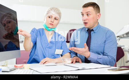Dentista femmina, puntando al monitor, spiegando diacrisis a sconvolto l uomo in studio dentistico Foto Stock