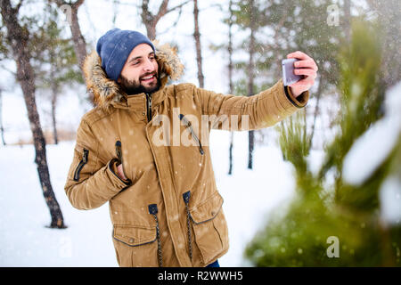 Sorridente uomo barbuto indossa caldo abbigliamento invernale e tenendo selfie foto con la fotocamera dello smartphone. Uomo bello scrivere messaggi con il cellulare e utilizzando le app in boschi innevati. Nevicata nel bosco. Foto Stock