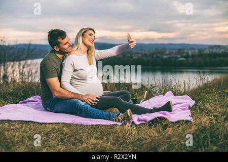 Felice il marito e la moglie incinta prendendo selfie mentre essi amano trascorrere del tempo insieme all'aperto. Foto Stock