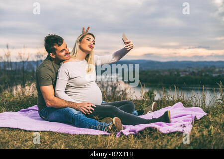 Felice il marito e la moglie incinta prendendo selfie mentre essi amano trascorrere del tempo insieme all'aperto. Foto Stock