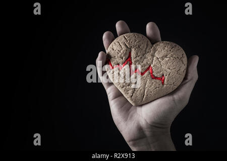 Primo piano della mano di un uomo caucasico tenendo a casa a forma di cuore, cookie con un elettrocardiogramma rosso in essa, contro uno sfondo nero con tom Foto Stock