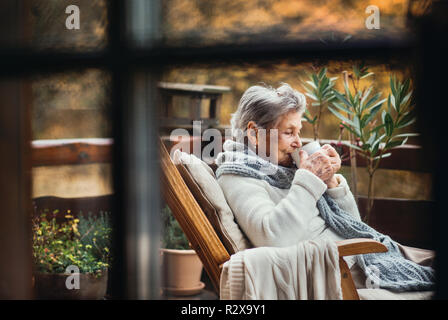 Una donna anziana seduti all'aperto su una terrazza in un giorno in autunno, bere caffè. Foto Stock