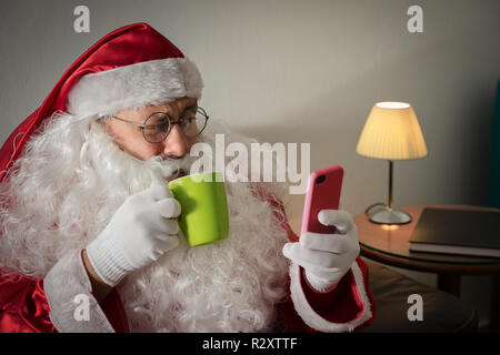 Profilo di metà di Santa Claus (o Babbo Natale) rilassante nel divano nel soggiorno utilizzando il dispositivo wireless on-line. Guardando al telefono cellulare e schermo Foto Stock