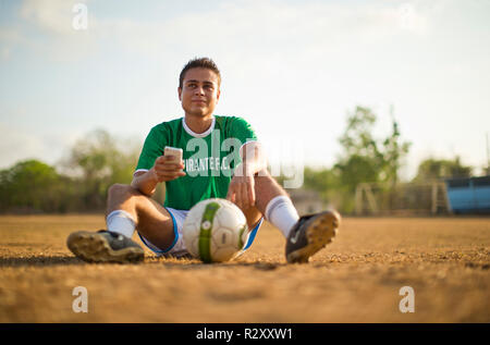 Contemplativo giovane giocatore di calcio di inviare un messaggio di testo mentre è seduto su un campo da gioco con un pallone da calcio. Foto Stock
