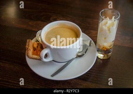 Tazza di caffè con un piccolo pezzo di torta di frutta e un trasparente aggiuntivo con il liquore e la panna montata Foto Stock