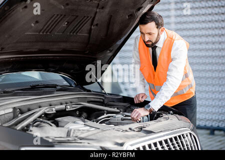 Imprenditore nel corpetto guardando al motore della sua Broken auto sulla strada Foto Stock