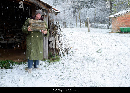 Uomo che porta legna da ardere attraverso la caduta di neve in giardino per uso come combustibile in un bruciatore di legno Zala county Ungheria Foto Stock