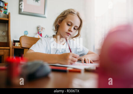 Una bambina di sette anni è seduto a casa a un tavolo e scrive in un notebook, il completamento di un compito di apprendimento o lezioni di ripetizione Foto Stock