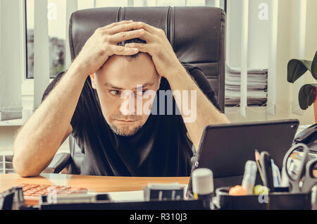 Scioccato imprenditore seduto in una sedia alla sua scrivania e guardando una schermata di tablet Foto Stock