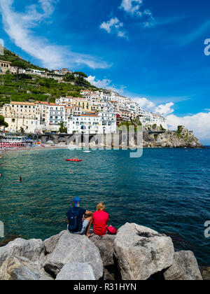 Giovane si siede sulla roccia e guarda la città vecchia e la Spiaggia di Amalfi, penisola di Sorrento, Costiera Amalfitana, Campania, Italia Foto Stock