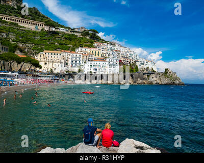 Giovane si siede sulla roccia e guarda la città vecchia e la Spiaggia di Amalfi, penisola di Sorrento, Costiera Amalfitana, Campania, Italia Foto Stock