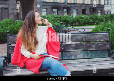 Un attraente ragazza con capelli lunghi marrone si siede su una panca, nascondendo dietro un tappeto rosso, rosicchia una penna e di pensiero su un background urbano. Essa dispone di un notebook nelle sue mani Foto Stock