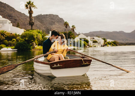 Coppia giovane seduti insieme in una barca di toccare le loro teste. Giovane avendo divertimento su una barca data in un lago con le colline e le case in background. Foto Stock