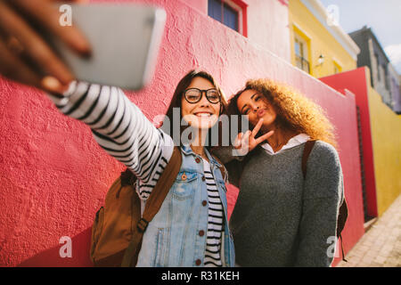 Due belle giovani donne di scattare una foto insieme. Giovani femmine prendendo un selfie utilizzando il telefono cellulare e altri facendo la pace segno a mano, all'esterno. Foto Stock