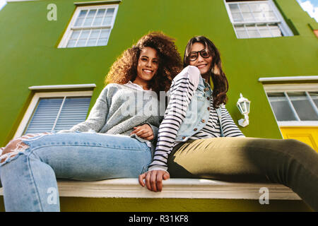 Basso angolo vista di due donne seduta sul banco di lavoro al di fuori di una casa verde. Amici di sesso femminile appendere fuori all'esterno guardando la fotocamera. Foto Stock