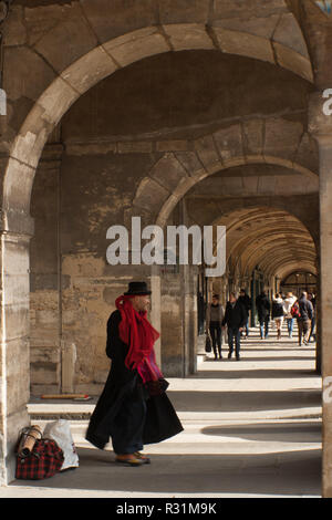 Artista di strada a Le Marais, Parigi, in piedi sotto archi di pietra, arcade, che indossa abiti neri e cappello e una sciarpa rossa. Punto di fuga Foto Stock