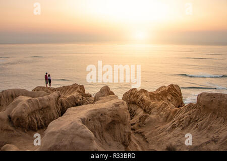 Due persone stanno nella distanza guardando verso un tramonto sull'oceano. Interessanti le scogliere e rocce portano lo sguardo verso il giovane e verso th Foto Stock