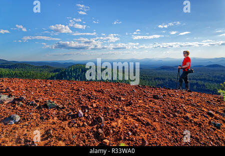 Donna 42,882.02712 escursionismo Alto di ampia bellissimo rosso vulcanico cono di scorie, alberi colline e boschi di conifere in distanza, cielo blu, il bianco delle nuvole Foto Stock