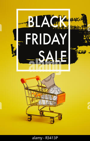 Vista ravvicinata del piccolo carrello della spesa con gli abiti di carta su sfondo giallo, venerdì nero vendita iscrizione Foto Stock