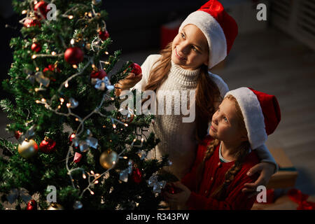 Due piccole sorelle di Santa caps guardando decorate albero di natale nella santa notte Foto Stock