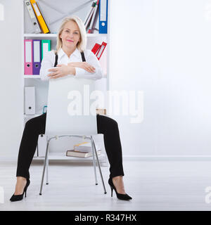 Bella bionda di mezza età della donna con sorriso raggiante seduta in ufficio guardando la telecamera Foto Stock