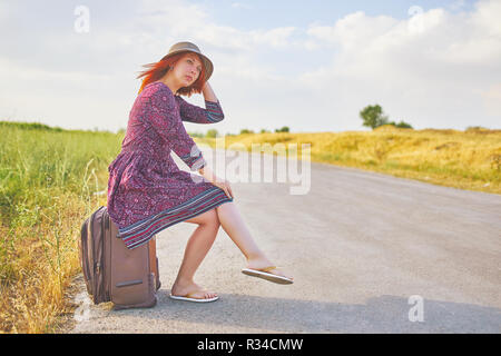 Molto bella donna in abiti estivi e hat autostop con la valigia sulla strada. Bellezza Donna seduta su bagagli in attesa di bus. Concetto di Hitchhiker Foto Stock