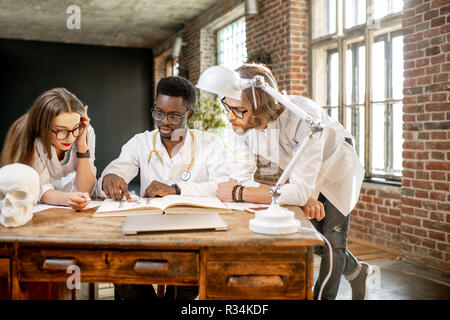 Gruppo di una giovane la multietnicità medici o di studenti di medicina in uniforme lavora con libro e disegni sul tavolo in ufficio o a scuola Foto Stock