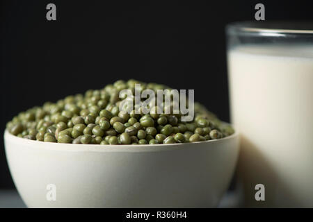 Primo piano di una ciotola con semi di soia e un bicchiere di latte di soia sul grigio di una tavola in legno rustico, contro uno sfondo nero Foto Stock