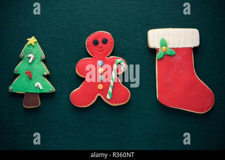 Alcuni biscotti fatti in casa con forme diverse come ad esempio un albero di Natale, un omino di pan di zenzero e una calza di Natale su uno sfondo verde scuro Foto Stock