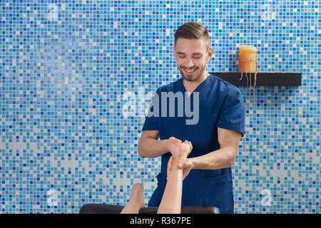 Vista frontale del maschio bello dottore in uniforme blu sorridente e parlare mentre facendo il massaggio dei piedi nella spa cosmetologia salon. Massaggiatore nel processo di lavorazione. Concetto di trattamento e cura. Foto Stock