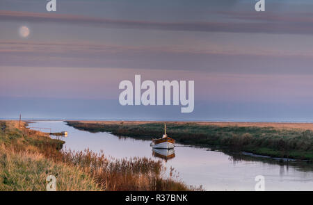 La luna piena che si innalzano per oltre Saltfleet Haven, Lincolnshire durante un tramonto in inverno, illuminando una solitaria barca sul fiume di marea a marea alta. Foto Stock