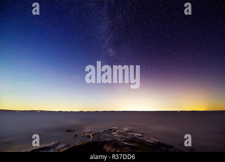 La Via Lattea e stelle nel tranquillo lago di scena a notte. Alcuni inquinamento luminoso e luci della città all'orizzonte Foto Stock