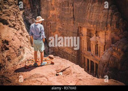 Turistico con cappello per il sole sorge su rocce e guarda dall'alto nella gola Siq, faraone treasure house scavata nella roccia, facciata Foto Stock