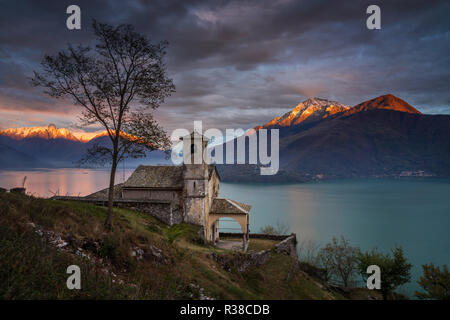 Chiesetta di Santa Eufemia a Musso oltre il Lago di Como con le montagne nella luce del tramonto sullo sfondo, Italia Foto Stock