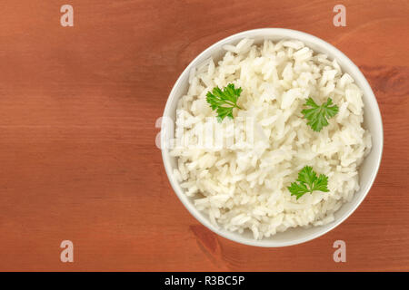 Una ciotola di cotto bianco a grani lunghi Riso Basmati, guarnite con foglie di prezzemolo, ripresa dall'alto su un rustico in legno con sfondo spazio copia Foto Stock