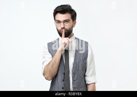 Ispanico maschio barbuto in round eyewear, mantiene il dito sulle labbra, chiede di non raccontare le informazioni segrete Foto Stock