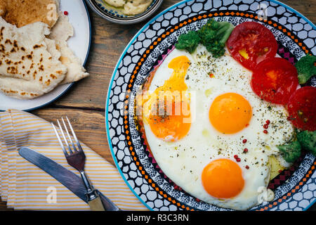 Tavolo per la colazione. Uova fritte su una piastra con pomodoro, pane pita) e hummus su un tavolo di legno. Vista dall'alto. Sfondo di legno. Foto Stock