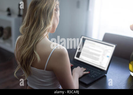 Donna bionda blogger scrivendo sul portatile a casa blog, vista posteriore Foto Stock