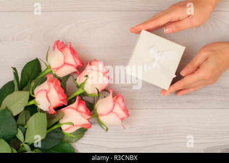 Donna di mani tenendo una confezione regalo su grigio Sfondo di legno con bellissime rose. Concetto di dare un regalo per le vacanze. Vista dall'alto. Foto Stock