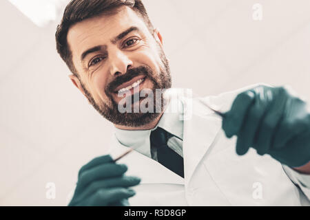 Allegro dentista. Close up di sorridere barbuto medico azienda strumenti odontoiatrici in mani e guardando la telecamera che esprimono ottimismo sul suo volto Foto Stock